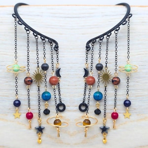 May include: Black ear cuffs with dangling chains and celestial charms. Each cuff has chains with beads in blue, red, and gold, plus star, moon, and planet charms. The design includes a sun and planet-shaped elements.