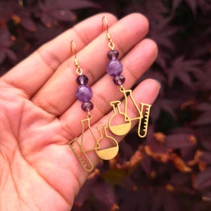 May include: A pair of gold-toned dangle earrings featuring laboratory glassware charms, including a beaker, a round-bottom flask, and a test tube. Each earring is accented with two purple spherical beads and one smaller faceted purple bead.