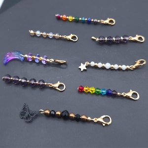 May include: A selection of beaded charms with gold-coloured clasps. The charms feature diverse designs, including a rainbow of beads, a star, a butterfly, and a crescent moon. The beads are in various colours, including clear, purple, and black.