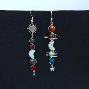 May include: A pair of mismatched silver-tone dangle earrings. One earring features a sun charm above a vertical arrangement of spherical beads in gray, red, and blue, and a white crescent moon. The other features wrapped wire, spherical beads in brown, yellow, green, and blue, a white crescent moon, and a star charm.