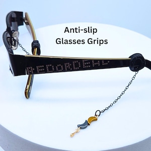 May include: Black and gold eyeglasses with a chain and decorative charm. The glasses have the text "REDOX EYEWEAR" on the arm. The image also includes the text "Anti-slip Glasses Grips".