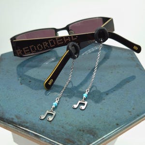 May include: Black sunglasses with gold trim and the text "#EDOROBE". Silver chains with musical note charms and turquoise beads hang from the frames. The sunglasses are displayed on a blue tile surface.