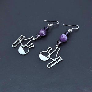 May include: A pair of silver-toned drop earrings featuring science-themed charms including a test tube, an Erlenmeyer flask, and a round-bottom flask. Each earring is accented with two dark purple beads and one lighter purple marbled bead.