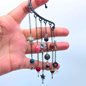 May include: A black metal ear cuff featuring multiple dangling chains adorned with various colorful spherical beads, a silver-toned sun charm, a silver-toned star charm, and two planets with silver wire rings encircling the beads.