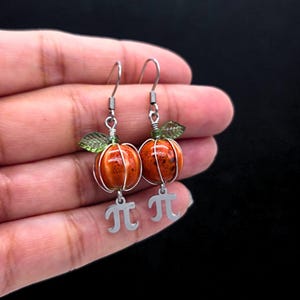 May include: A pair of orange pumpkin earrings with silver wire detailing and green leaf accents. Each earring has a silver charm with the mathematical symbol pi. The earrings are displayed against a dark background.