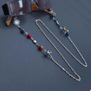 May include: Silver-coloured eyeglass chains with a celestial theme. The chains feature colourful beads, stars, and planet charms. One chain has a sun charm. The eyeglasses are black.