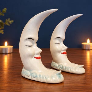 May include: A pair of ceramic bookends shaped like crescent moons with closed eyes and red painted lips. The moons are white with a light blue cloud-like base. Each moon features a serene facial expression and a smooth, glossy finish.