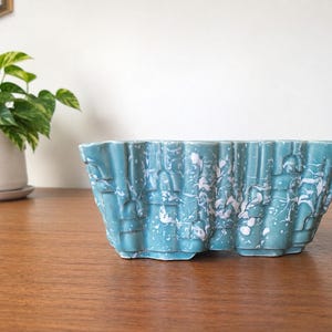 May include: A rectangular ceramic planter with a teal blue base color and a white speckled, marbled finish. The exterior features a repeating vertical ribbed or pleated texture that creates a series of recessed and raised geometric panels.