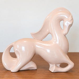 May include: A pale pink ceramic horse figurine in a stylized, reclining pose. The sculpture features a smooth, glossy finish with flowing, curved lines that define the mane, tail, and body, creating a continuous, fluid silhouette throughout.