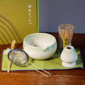 May include: A matcha tea set with a green and white ceramic bowl, a bamboo whisk, a metal sieve, and a bamboo scoop. The set includes a green and white whisk holder and a box with Japanese text.