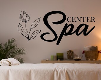 May include: A white spa treatment table with a rolled towel on top. The wall behind the table has a black and white floral design and the text "Spa Center" in black.