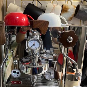 Pode incluir: M&aacute;quina de caf&eacute; expresso em a&ccedil;o inoxid&aacute;vel com detalhes em madeira e um man&oacute;metro. Uma caneca vermelha e outras canecas s&atilde;o armazenadas acima da m&aacute;quina. A m&aacute;quina tem um visor digital e uma vareta de vapor. A marca "Lelit" &eacute; vis&iacute;vel.