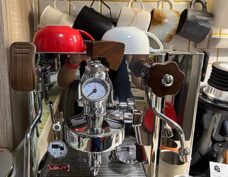Pode incluir: M&aacute;quina de caf&eacute; expresso em a&ccedil;o inoxid&aacute;vel com detalhes em madeira e um man&oacute;metro. Uma caneca vermelha e outras canecas s&atilde;o armazenadas acima da m&aacute;quina. A m&aacute;quina tem um visor digital e uma vareta de vapor. A marca "Lelit" &eacute; vis&iacute;vel.