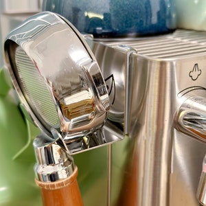 Puede incluir: Un portafiltro de espresso cromado con mango de madera, fijado a una máquina de espresso de acero inoxidable.