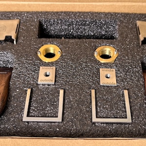 Pode incluir: Um kit de componentes de hardware, incluindo puxadores de madeira, suportes de metal e acess&oacute;rios de lat&atilde;o, dispostos em uma caixa de papel&atilde;o com inser&ccedil;&otilde;es de espuma. As pe&ccedil;as de metal s&atilde;o prateadas e douradas, algumas com o texto "inside" e "outside".