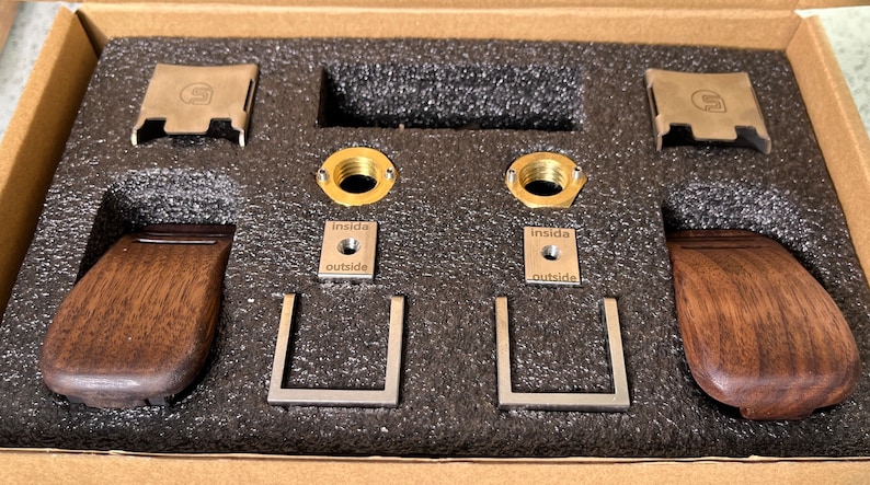 Pode incluir: Um kit de componentes de hardware, incluindo puxadores de madeira, suportes de metal e acess&oacute;rios de lat&atilde;o, dispostos em uma caixa de papel&atilde;o com inser&ccedil;&otilde;es de espuma. As pe&ccedil;as de metal s&atilde;o prateadas e douradas, algumas com o texto "inside" e "outside".