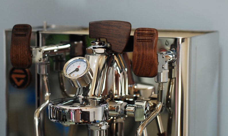 Pode incluir: Close-up de uma m&aacute;quina de caf&eacute; expresso em a&ccedil;o inoxid&aacute;vel com detalhes em madeira. Apresenta um man&oacute;metro, pegas de madeira e v&aacute;rios componentes cromados. A m&aacute;quina tem uma superf&iacute;cie polida e refletora.