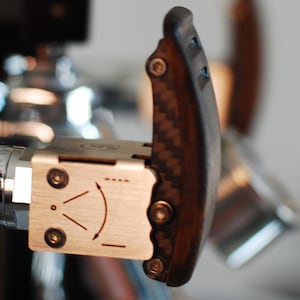 Pode incluir: Close-up de uma alavanca de m&aacute;quina de caf&eacute; com uma pega em madeira escura e fibra de carbono. A alavanca est&aacute; presa a um componente de a&ccedil;o inoxid&aacute;vel com um indicador direcional. A pega &eacute; curva e ergon&oacute;mica.