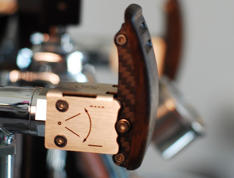 Pode incluir: Close-up de uma alavanca de m&aacute;quina de caf&eacute; com uma pega em madeira escura e fibra de carbono. A alavanca est&aacute; presa a um componente de a&ccedil;o inoxid&aacute;vel com um indicador direcional. A pega &eacute; curva e ergon&oacute;mica.