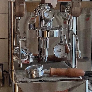 Pode incluir: M&aacute;quina de caf&eacute; expresso em a&ccedil;o inoxid&aacute;vel com detalhes em madeira. Possui um porta-filtro, man&oacute;metros e um aquecedor de ch&aacute;venas. A m&aacute;quina, com acabamento cromado, assenta numa base de madeira. A palavra "ELIT" &eacute; vis&iacute;vel.