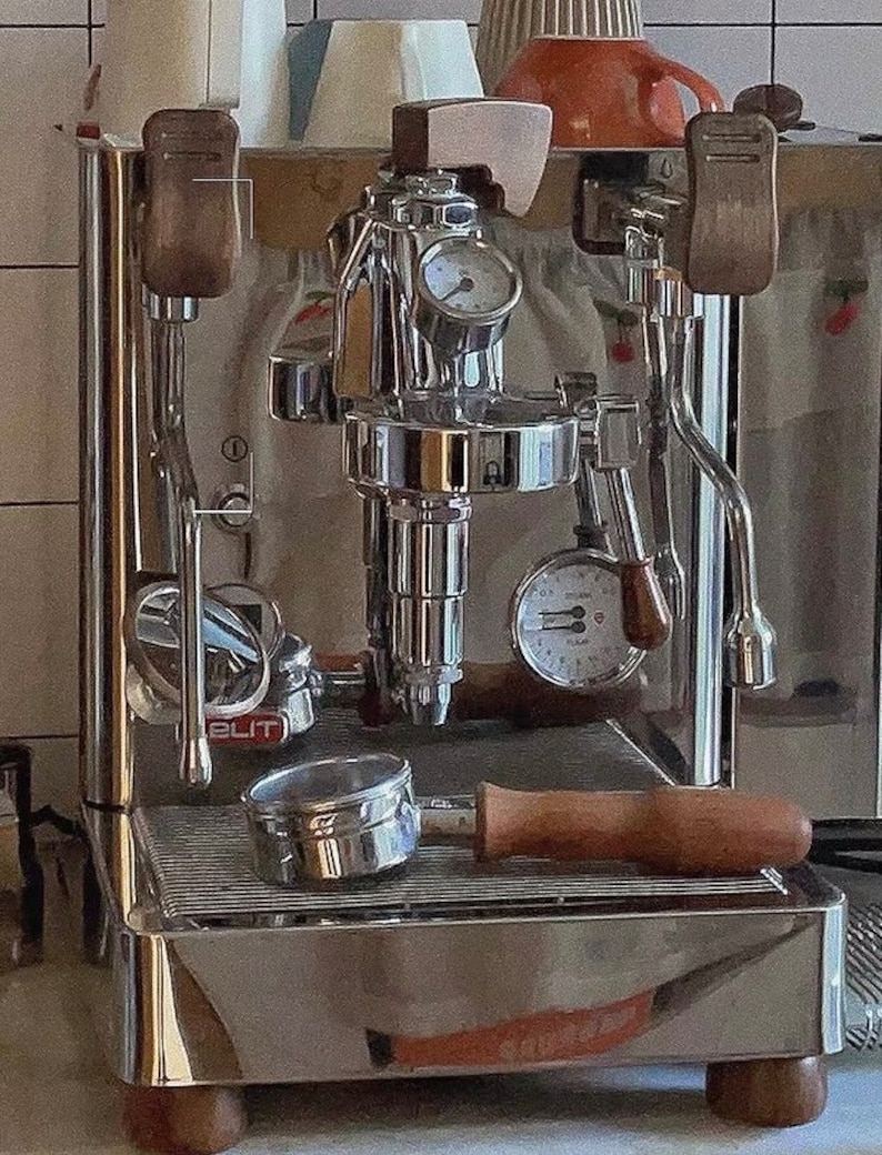 Pode incluir: M&aacute;quina de caf&eacute; expresso em a&ccedil;o inoxid&aacute;vel com detalhes em madeira. Possui um porta-filtro, man&oacute;metros e um aquecedor de ch&aacute;venas. A m&aacute;quina, com acabamento cromado, assenta numa base de madeira. A palavra "ELIT" &eacute; vis&iacute;vel.