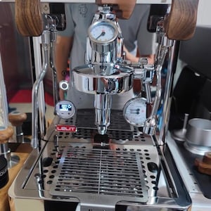 Pode incluir: M&aacute;quina de caf&eacute; expresso em a&ccedil;o inoxid&aacute;vel polido com detalhes em madeira. Possui um man&oacute;metro, um visor digital e uma bandeja de gotejamento. A marca "LELIT" &eacute; vis&iacute;vel. A m&aacute;quina tem uma superf&iacute;cie reflexiva.