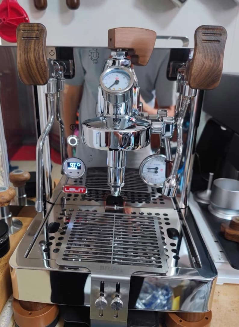 Pode incluir: M&aacute;quina de caf&eacute; expresso em a&ccedil;o inoxid&aacute;vel polido com detalhes em madeira. Possui um man&oacute;metro, um visor digital e uma bandeja de gotejamento. A marca "LELIT" &eacute; vis&iacute;vel. A m&aacute;quina tem uma superf&iacute;cie reflexiva.