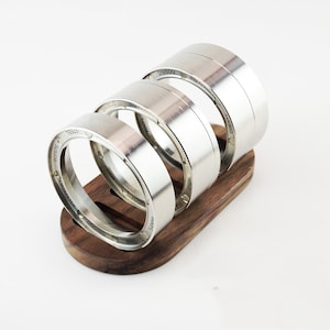 Op de afbeelding: Een set roestvrijstalen doseerringen voor een koffiemachine, gepresenteerd op een houten standaard. De ringen hebben gegraveerde tekst, waaronder "54MM", "DOSING RING" en "STAINLESS STEEL". De ringen zijn gestapeld, wat hun verschillende hoogtes laat zien.