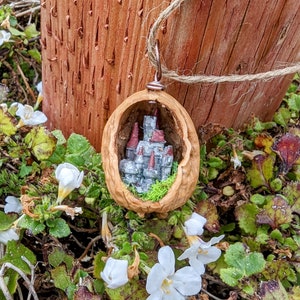 Puede incluir: Un colgante único con un castillo en miniatura dentro de una cáscara de nuez. El castillo tiene muros grises y torres con tejados rojos, sobre musgo verde. El colgante cuelga de un lazo color cobre y una cuerda natural.