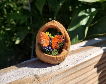 Può includere: Un diorama artigianale a guscio di noce con una vivace farfalla arancione con segni neri, immersa tra fiori e vegetazione in miniatura. La farfalla è il punto focale, incastonata nel guscio naturale, creando un pezzo decorativo unico.
