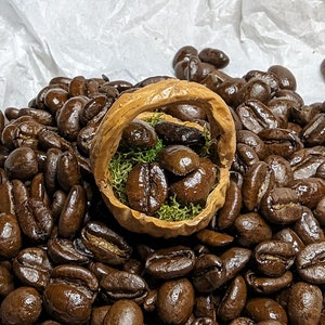 May include: A close-up shot of roasted coffee beans, with a small, decorative basket filled with coffee beans and green moss. The basket is made of a light brown material and sits amidst a pile of dark brown coffee beans.