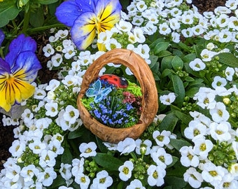 Può includere: Un piccolo cestino di legno con una scena di giardino in miniatura, tra cui una coccinella e una farfalla, è circondato da fiori bianchi, foglie verdi e viole blu e gialle, creando un affascinante giardino in miniatura.