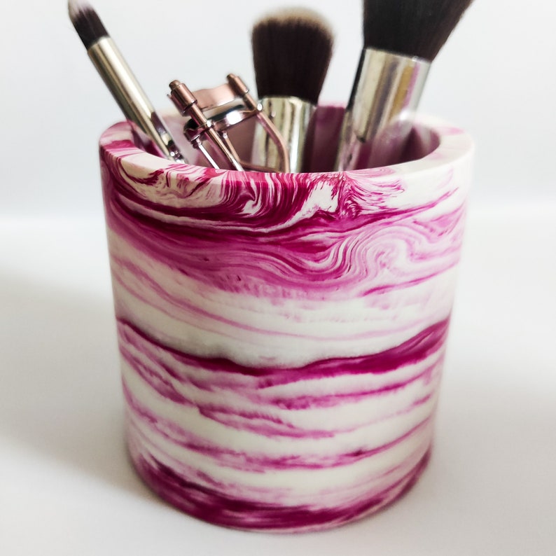May include: A pink and white marbled ceramic makeup brush holder with a variety of brushes and a lash curler inside.