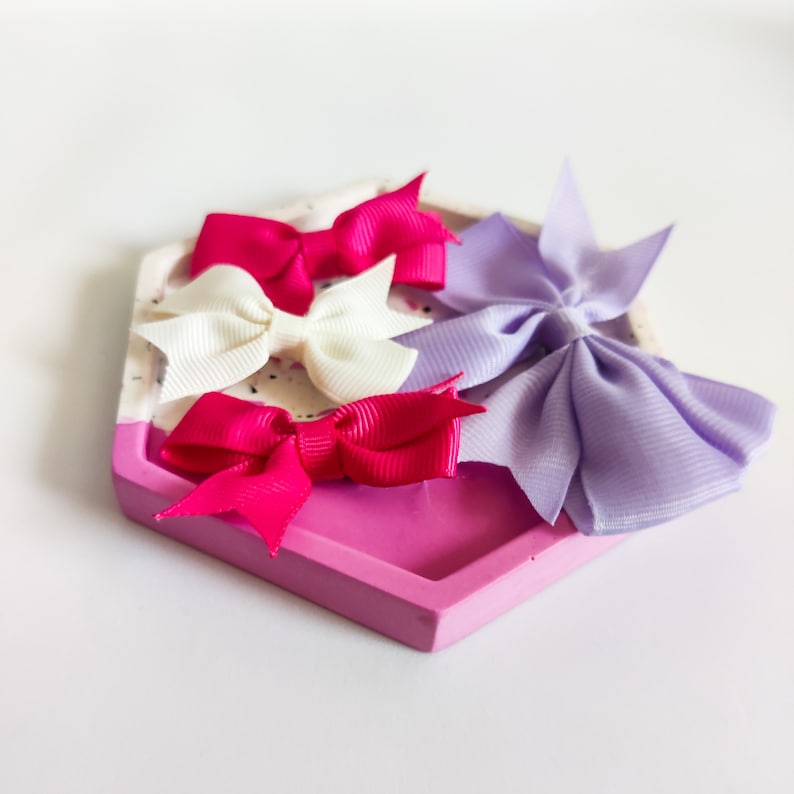 May include: Three hair bows in a pink and white hexagonal tray. The bows are pink, white, and lavender.