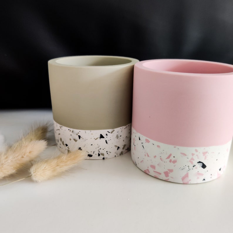 Two cylindrical planters/pen pots, one in a light sage green color and the other in pink. Both pen pots have a white base with black and pink/sage green speckled pattern around the bottom edge.