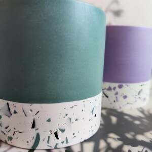 Two cylindrical planters, one teal and one purple, with a white base and a speckled pattern. The planters are made of a smooth material.