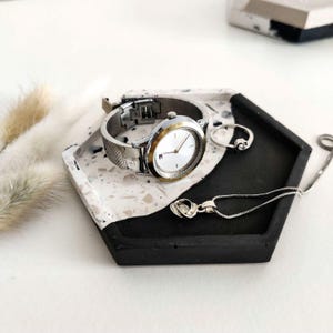 May include: A black and white hexagonal tray with a silver watch and a silver necklace with a heart pendant. The tray is made of concrete and has a speckled finish.