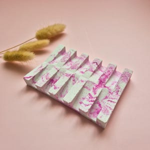 May include: A white soap dish with pink marbling. The dish has a rectangular shape with raised ridges for draining water.