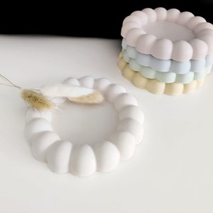 May include: A set of four white, light blue, light green, and yellow silicone coasters with a raised, rounded, flower-like design. The coasters are stacked on top of each other.