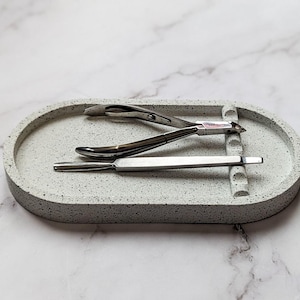 May include: A gray speckled concrete tray holds two silver nail tools. One is a curved cuticle nipper, and the other is a flat, pointed tool. The tray is oval-shaped.