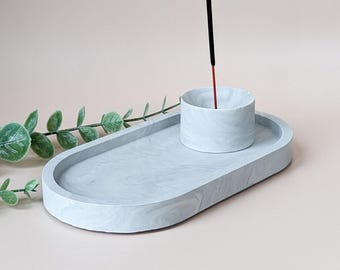 May include: A light gray incense holder set. The set includes an oval tray and a cylindrical incense stick holder. The holder has a marbled design. An incense stick with a red tip is in the holder. A green leafy branch is in the background.