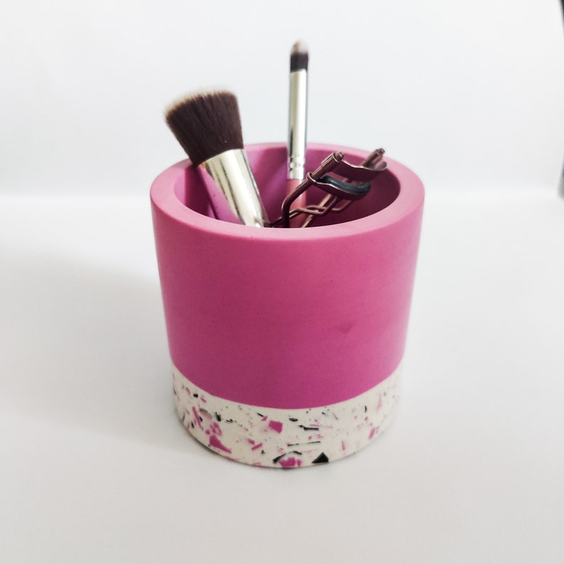 May include: A pink and white speckled ceramic makeup brush holder with a black and white speckled base. The holder contains a black and brown makeup brush, a silver eyelash curler, and a silver makeup brush.