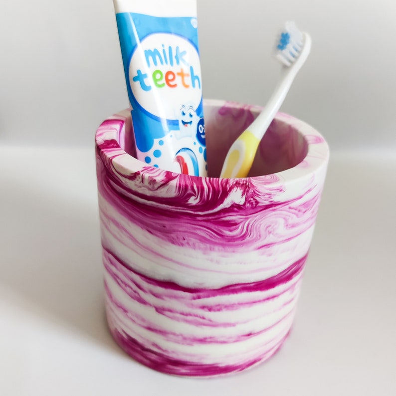 May include: A pink and white marbled ceramic toothbrush holder with a tube of "milk teeth" toothpaste and a toothbrush inside.