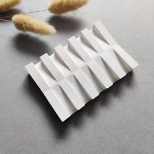 May include: A white, geometric soap dish with a textured surface. The dish features a series of angled, parallel ridges, creating a zig-zag pattern. The design is minimalist and modern, suitable for bathroom decor.