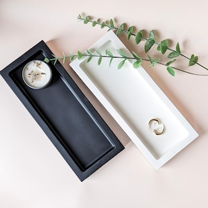 May include: Two rectangular trays, one black and one white, are displayed on a pink surface. The black tray holds a small candle, while the white tray contains a gold ring. Greenery adds a natural touch to the arrangement.
