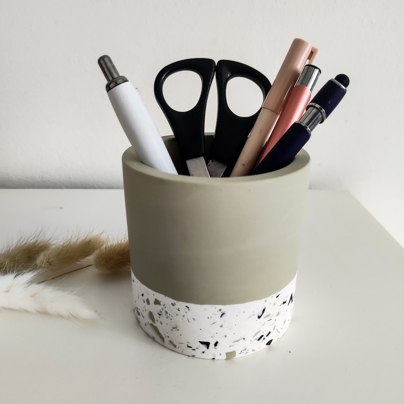 A sage green and white terrazzo pencil holder with a white pen, a pink pen, a black pen, a stylus, and a pair of black scissors inside.