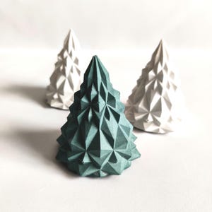 May include: Three geometric Christmas tree decorations. One is teal, and two are white. The trees have a faceted design, creating a modern, minimalist aesthetic. The trees are arranged on a white surface.