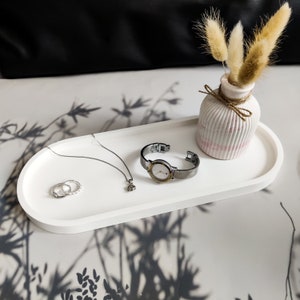May include: An oval white tray holding a silver-tone wristwatch with a round face, a delicate silver chain necklace with a small pendant, two silver rings, and a small ribbed white vase containing three dried beige grass plumes.