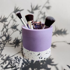A purple and white speckled jesmonite makeup brush holder with three makeup brushes and a lash curler inside