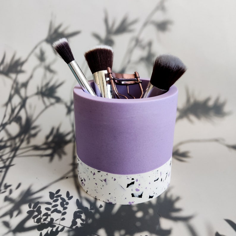 A purple and white speckled jesmonite makeup brush holder with three makeup brushes and a lash curler inside